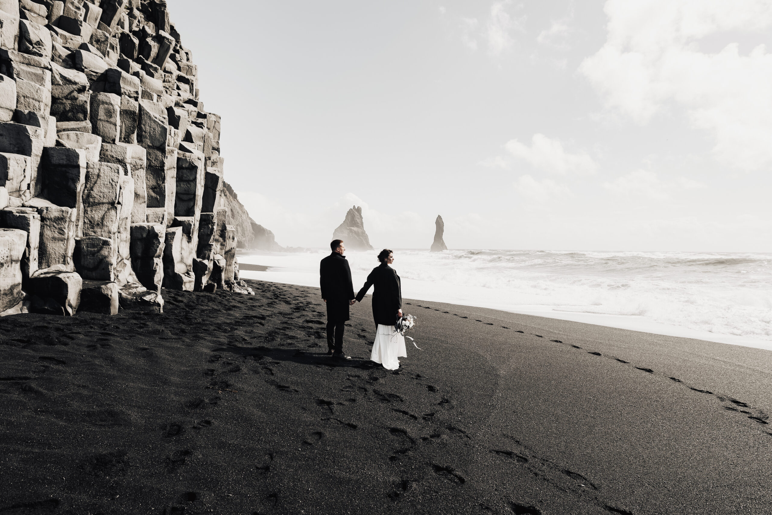 The wedding couple walks along the sandy black beach Vik, near the basalt cliff, in the form of pillars. The bride and groom hold hands and walk along the shore.